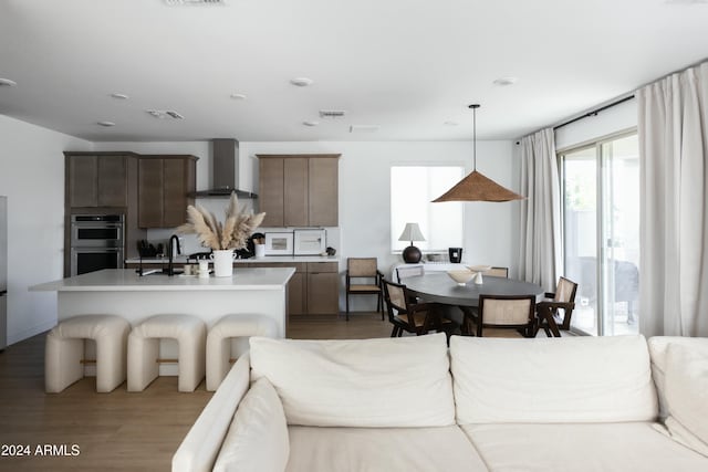 kitchen featuring light hardwood / wood-style flooring, an island with sink, stainless steel double oven, wall chimney range hood, and pendant lighting