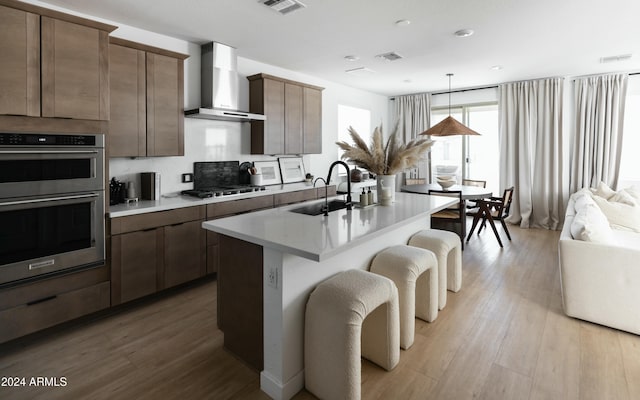 kitchen with a center island with sink, appliances with stainless steel finishes, sink, light hardwood / wood-style flooring, and wall chimney range hood