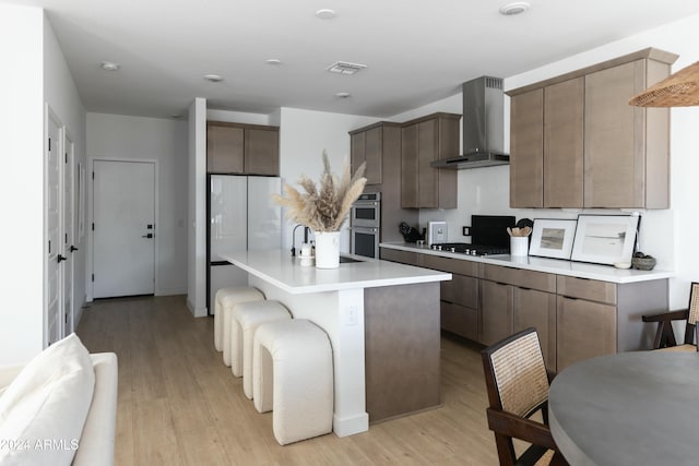 kitchen featuring wall chimney exhaust hood, stainless steel appliances, light hardwood / wood-style floors, a breakfast bar, and a kitchen island