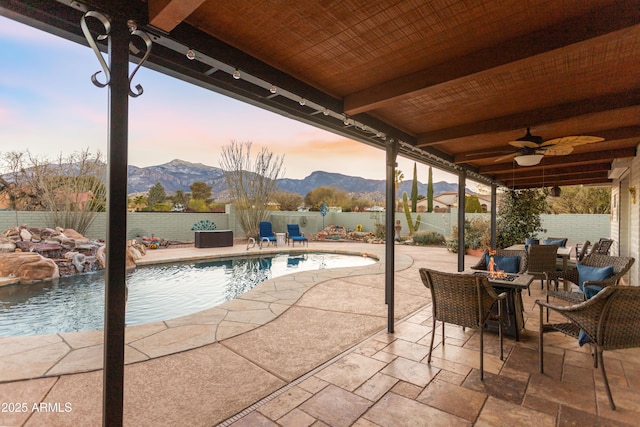 patio terrace at dusk featuring pool water feature, a mountain view, ceiling fan, and a fenced in pool