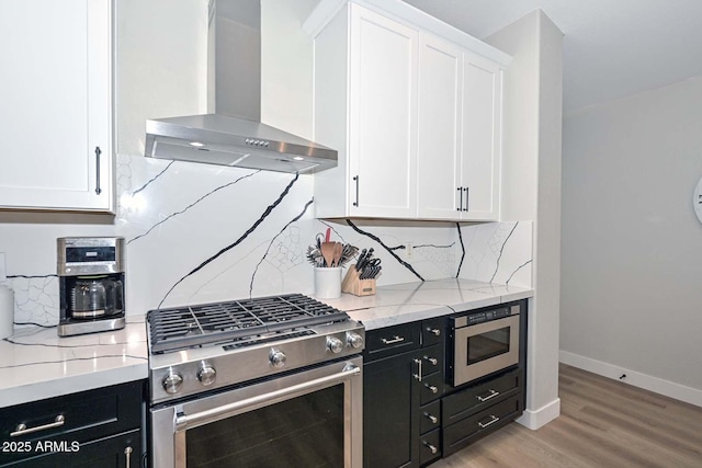 kitchen with appliances with stainless steel finishes, white cabinetry, light stone countertops, wall chimney exhaust hood, and light wood-type flooring