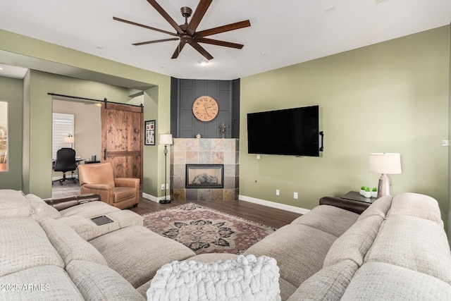 living room with ceiling fan, a barn door, baseboards, dark wood-style floors, and a tiled fireplace