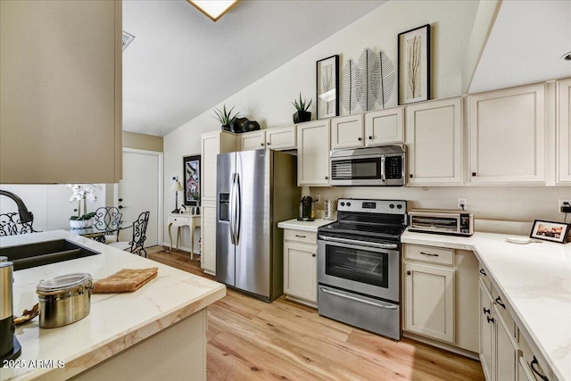 kitchen featuring stainless steel appliances, a sink, vaulted ceiling, light wood-style flooring, and light stone countertops