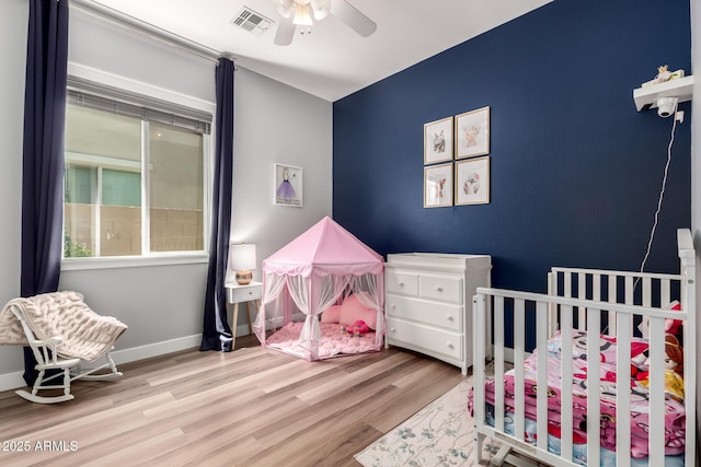 bedroom featuring visible vents, baseboards, a nursery area, wood finished floors, and a ceiling fan