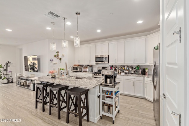 kitchen featuring stainless steel appliances, light stone counters, decorative light fixtures, a center island with sink, and white cabinets