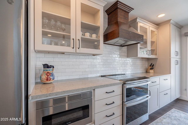 kitchen with dark wood-style flooring, light stone countertops, glass insert cabinets, wall chimney exhaust hood, and stainless steel appliances