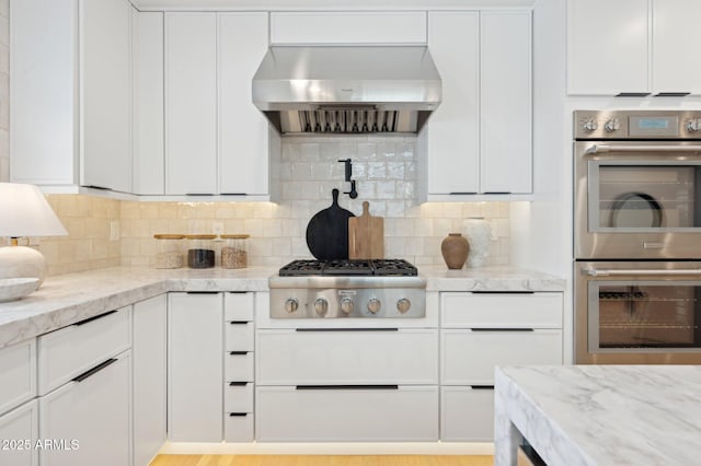kitchen with light stone counters, backsplash, appliances with stainless steel finishes, white cabinets, and wall chimney range hood
