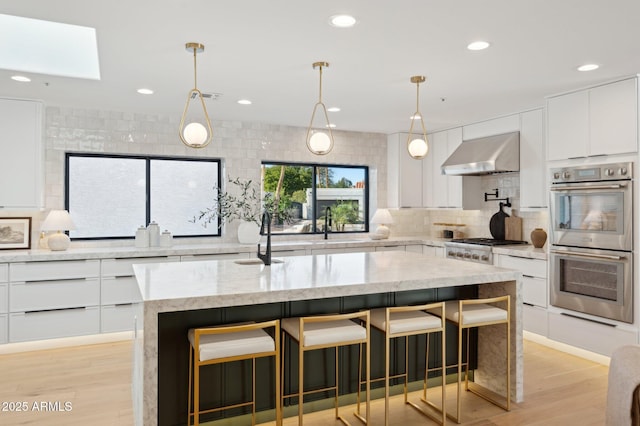 kitchen featuring stainless steel double oven, light wood-style flooring, decorative backsplash, white cabinets, and wall chimney exhaust hood