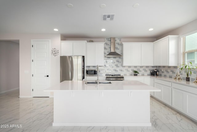 kitchen with stainless steel appliances, white cabinetry, a kitchen island with sink, and wall chimney range hood