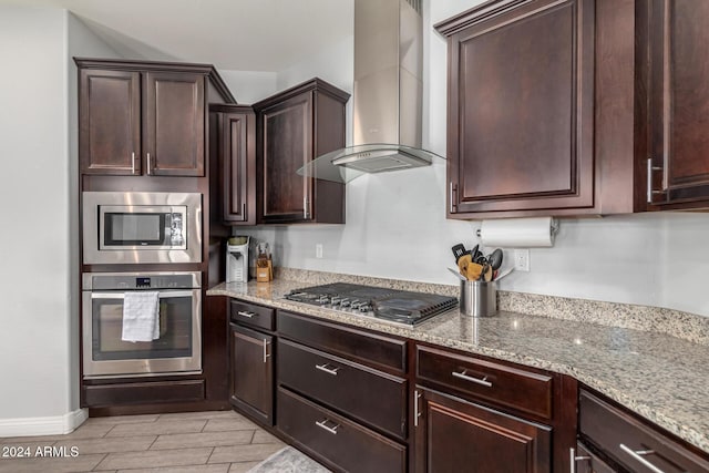 kitchen featuring dark brown cabinetry, wall chimney range hood, light stone counters, light hardwood / wood-style flooring, and appliances with stainless steel finishes