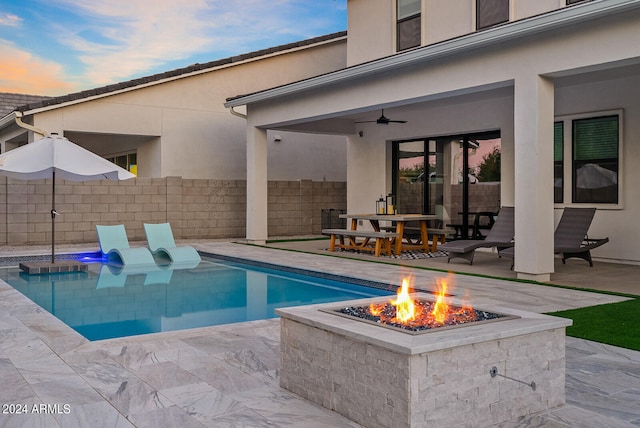 pool at dusk with ceiling fan, a patio area, and an outdoor fire pit