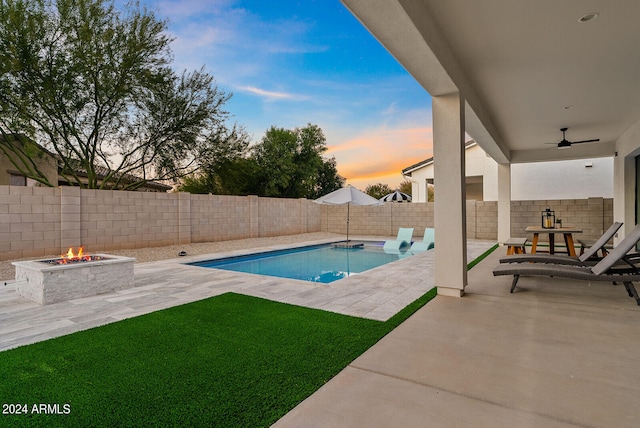 pool at dusk with ceiling fan, a fire pit, a lawn, and a patio