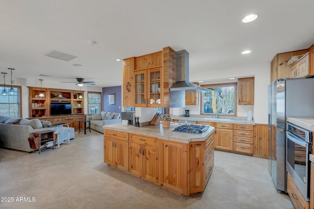 kitchen with ceiling fan, appliances with stainless steel finishes, a center island, tile counters, and island exhaust hood