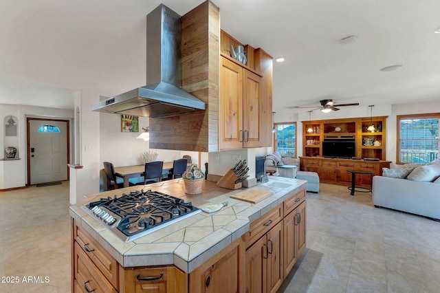 kitchen with sink, stainless steel gas cooktop, tile counters, ceiling fan, and wall chimney range hood