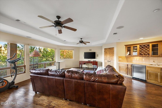 living room with dark wood-type flooring, ceiling fan, a tray ceiling, and wet bar