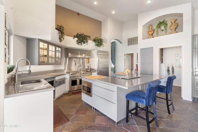 kitchen featuring built in appliances, a sink, visible vents, wall chimney range hood, and washing machine and clothes dryer