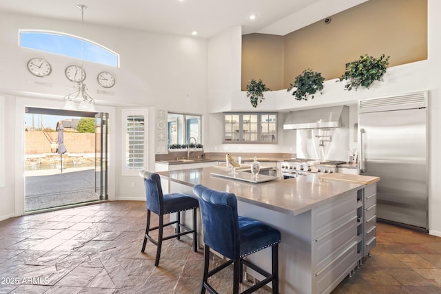 kitchen featuring high end appliances, a towering ceiling, wall chimney exhaust hood, stone tile flooring, and a sink