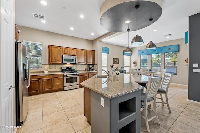 kitchen with pendant lighting, light tile patterned floors, appliances with stainless steel finishes, light stone countertops, and a center island with sink