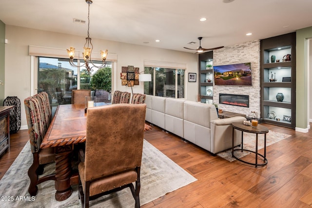 dining space featuring hardwood / wood-style floors, built in shelves, a stone fireplace, and ceiling fan with notable chandelier