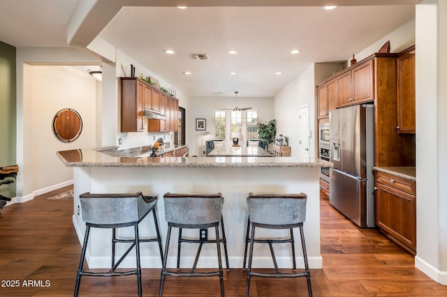 kitchen with appliances with stainless steel finishes, a breakfast bar area, dark hardwood / wood-style flooring, light stone counters, and kitchen peninsula