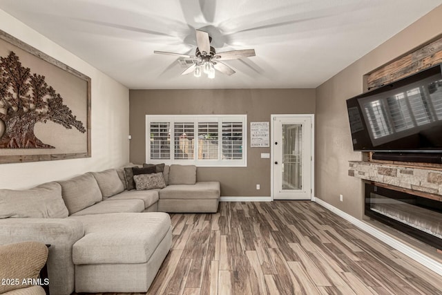 living room featuring wood finished floors, a ceiling fan, baseboards, and a stone fireplace