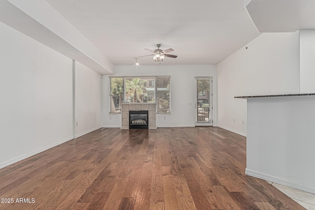 unfurnished living room with wood-type flooring, ceiling fan, a tiled fireplace, and baseboards