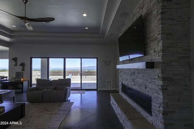 tiled living room with ceiling fan, a stone fireplace, and a tray ceiling