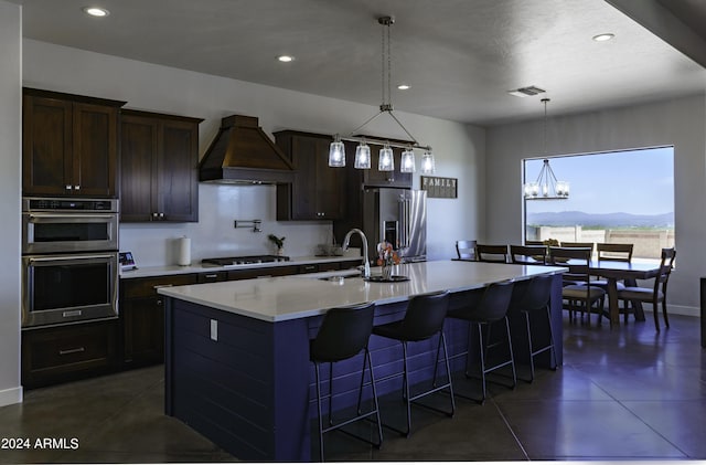 kitchen featuring appliances with stainless steel finishes, custom range hood, a kitchen island with sink, sink, and a chandelier