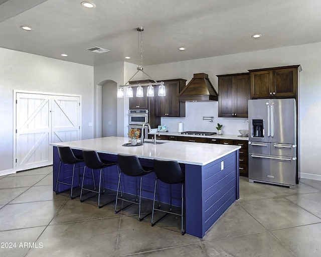 kitchen with custom exhaust hood, a center island with sink, hanging light fixtures, appliances with stainless steel finishes, and a kitchen bar