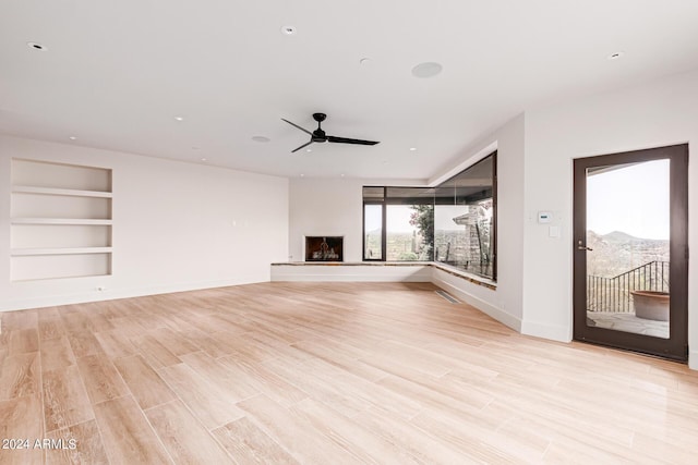 unfurnished living room with baseboards, a fireplace with raised hearth, a ceiling fan, light wood-style flooring, and built in shelves