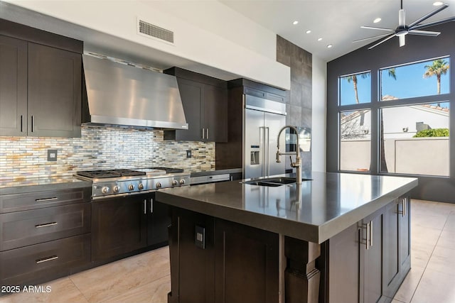 kitchen with wall chimney exhaust hood, stainless steel appliances, sink, backsplash, and a kitchen island with sink