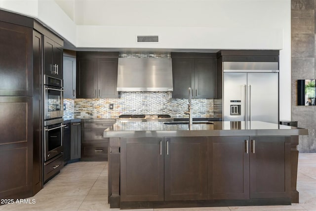 kitchen with an island with sink, sink, appliances with stainless steel finishes, and wall chimney range hood