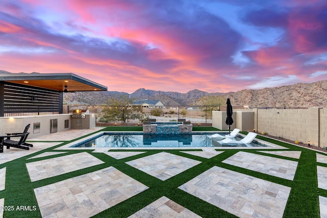 pool at dusk featuring an outdoor kitchen, a patio, a fenced backyard, a pool with connected hot tub, and a mountain view