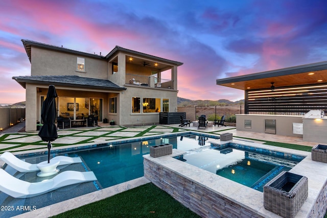 pool at dusk with a mountain view, fence, a pool with connected hot tub, a ceiling fan, and a patio area