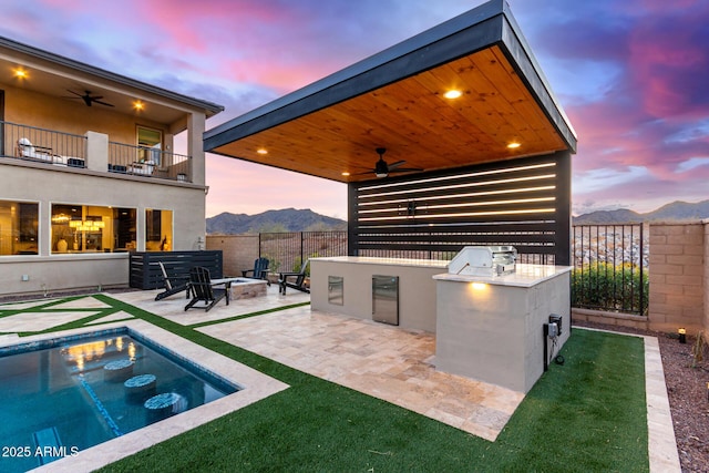 patio terrace at dusk featuring area for grilling, ceiling fan, a balcony, a mountain view, and a fire pit