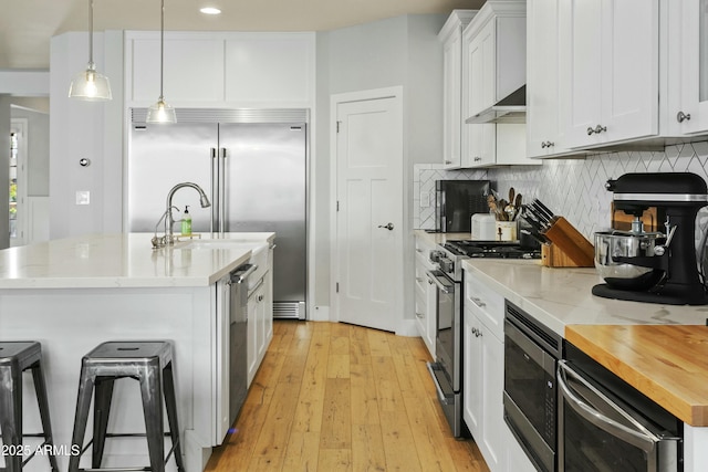 kitchen featuring built in appliances, pendant lighting, a center island with sink, and white cabinets