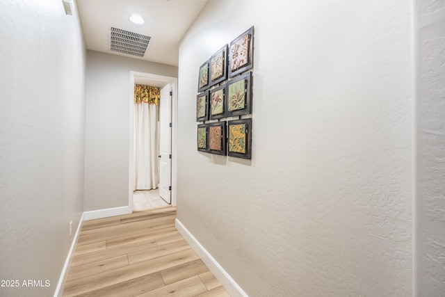 corridor with light wood-style flooring, visible vents, baseboards, and a textured wall