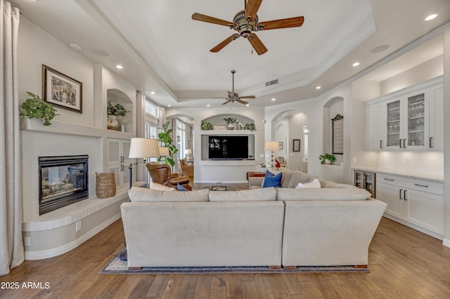 living room with built in shelves, a brick fireplace, a raised ceiling, ceiling fan, and light hardwood / wood-style floors