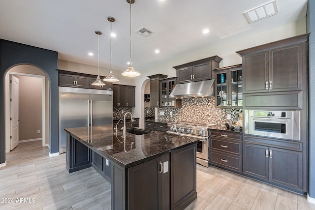 kitchen with high end appliances, sink, pendant lighting, dark stone countertops, and an island with sink