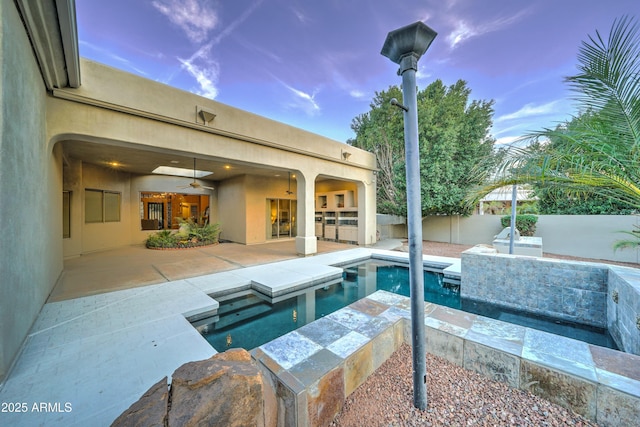 pool at dusk with exterior kitchen, a patio area, ceiling fan, and a jacuzzi