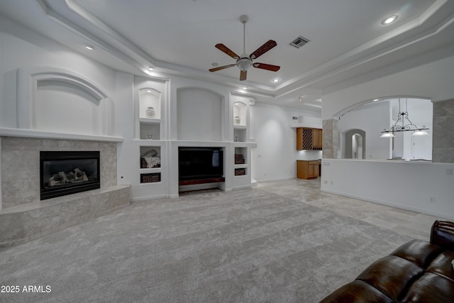 unfurnished living room featuring built in shelves, a raised ceiling, light colored carpet, a premium fireplace, and ceiling fan with notable chandelier