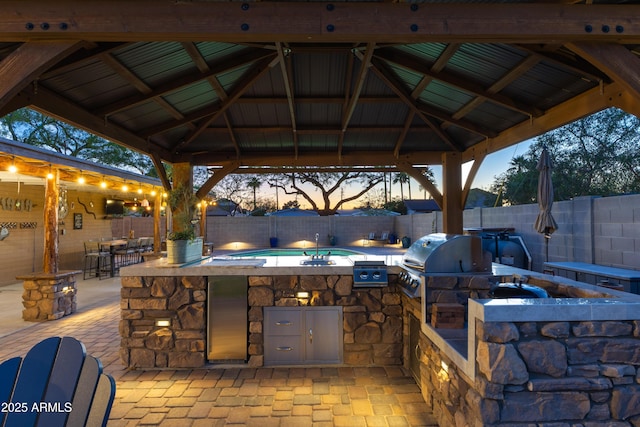 patio terrace at dusk featuring a fenced backyard, a gazebo, exterior kitchen, and a bar