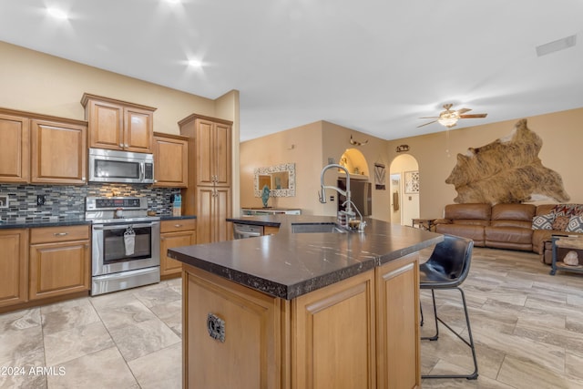 kitchen with backsplash, appliances with stainless steel finishes, an island with sink, sink, and ceiling fan