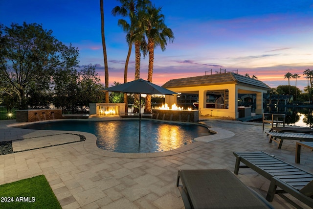 pool at dusk featuring a patio, pool water feature, a fire pit, and exterior kitchen