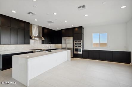 kitchen featuring visible vents, a kitchen island with sink, a sink, appliances with stainless steel finishes, and wall chimney range hood