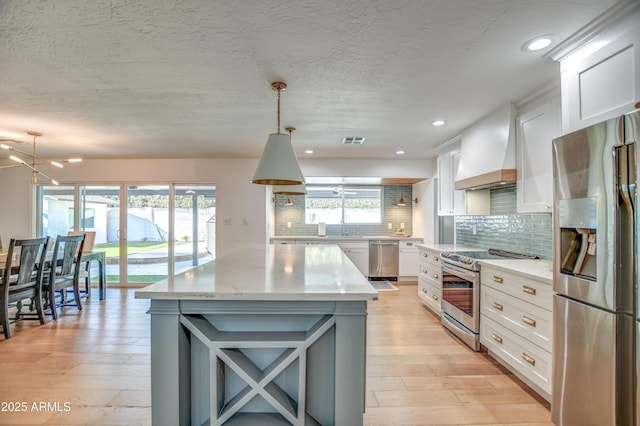 kitchen with pendant lighting, appliances with stainless steel finishes, white cabinetry, backsplash, and custom range hood