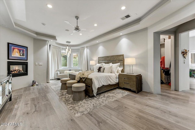 bedroom with a multi sided fireplace, light wood-type flooring, ceiling fan, and a raised ceiling