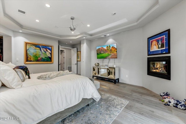 bedroom with light wood-type flooring, a multi sided fireplace, ceiling fan, and a raised ceiling