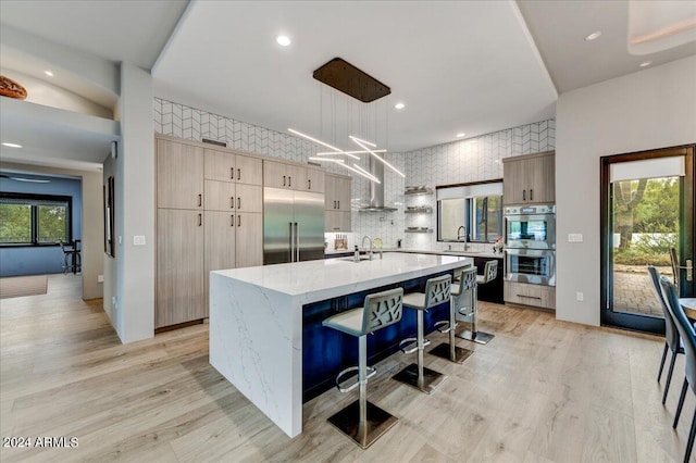 kitchen with hanging light fixtures, tasteful backsplash, a kitchen island with sink, light wood-type flooring, and appliances with stainless steel finishes