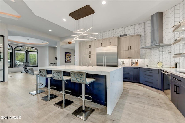 kitchen with a kitchen breakfast bar, an island with sink, decorative light fixtures, wall chimney range hood, and appliances with stainless steel finishes